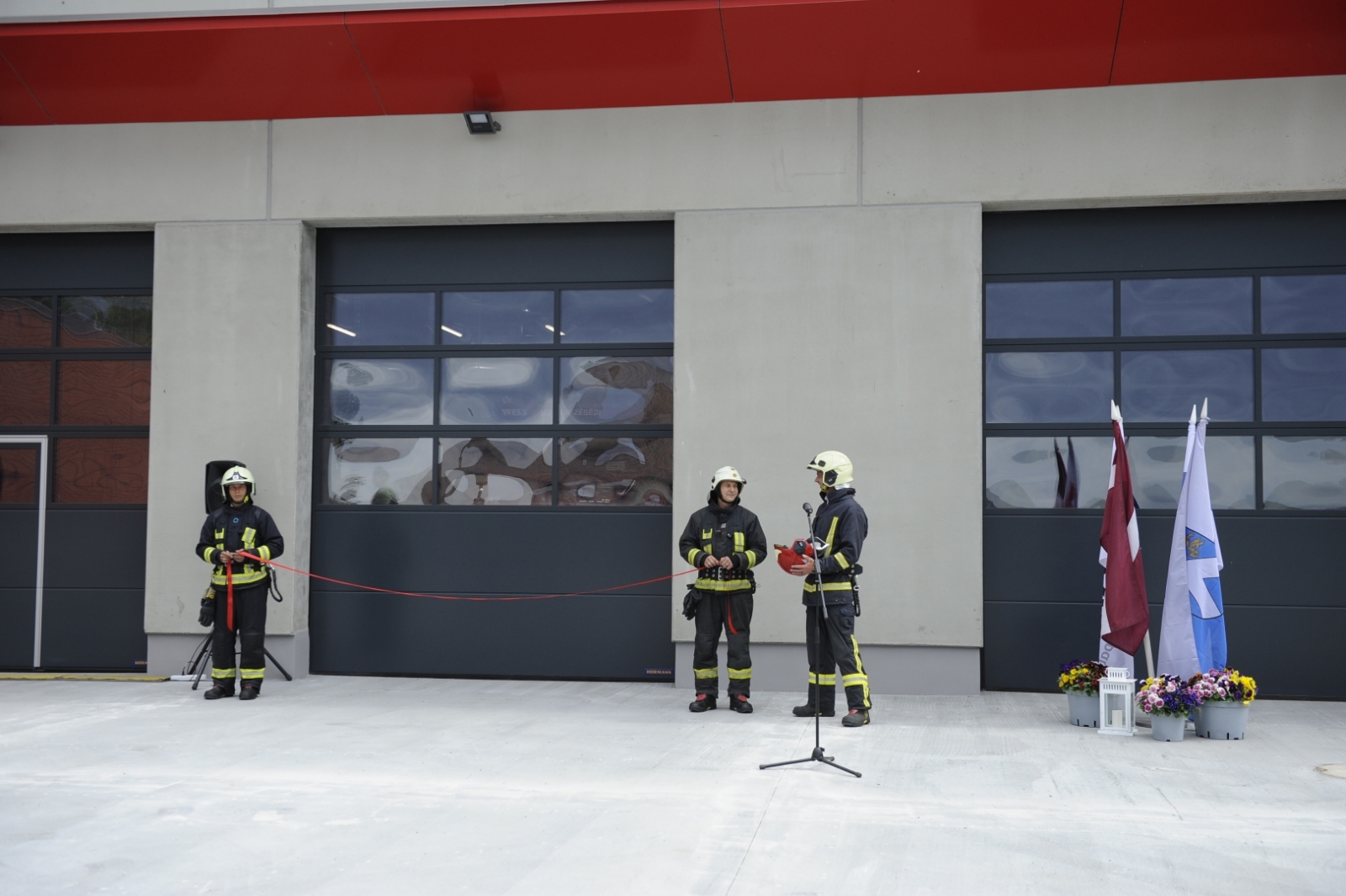Pie VUGD pelēki sarkanā DEPO stāv trīs ugunsdzēsēji glābēji. Divi no viņiem stāv katrs savā pusē ieejas durvīm, turot rokās sarkanu lenti.