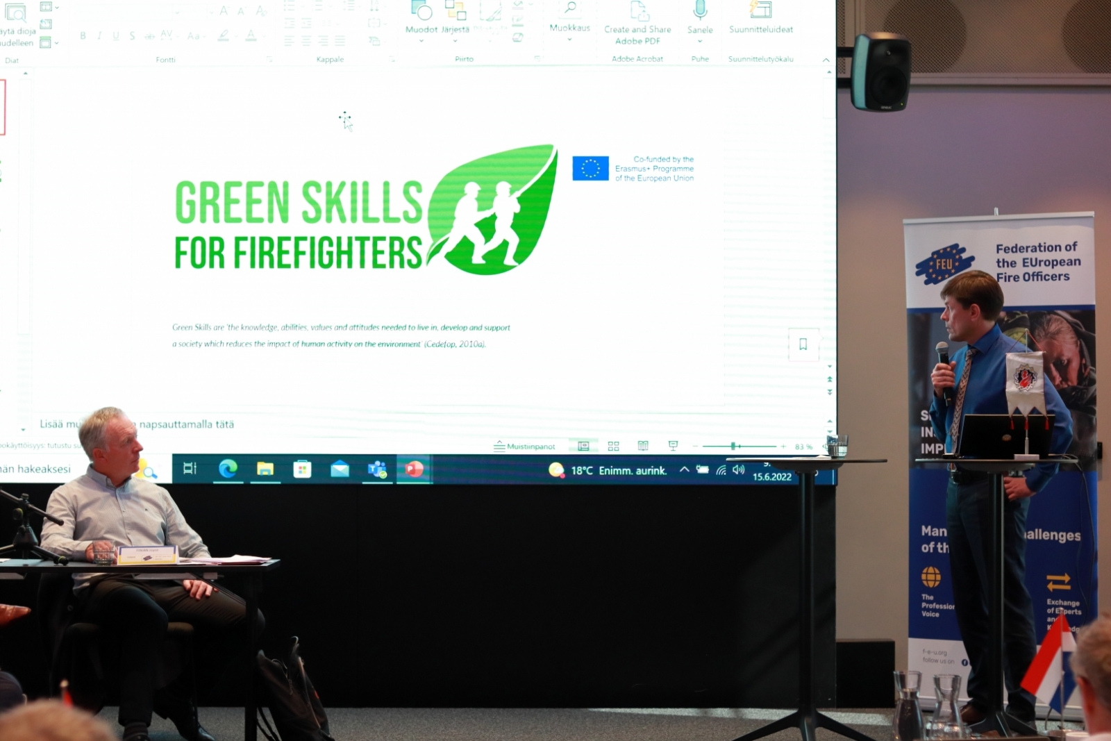 Auditorijas priekšā stāv vīrietis un rāda prezentāciju. Uz prezentācijas slaida redzams uzraksts angļu valodā "Green skills for firefighters"
