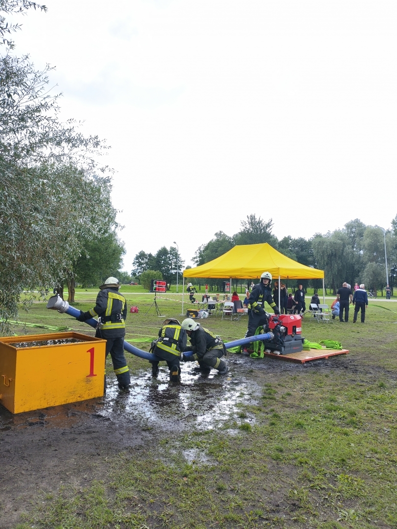 Trīs ugunsdzēsēji glābēji atrodas dubļainā zālienā. Viens stāv pie dzeltenas krāsas konteinera ar ūdeni. Divi ugunsdzēsēji pieliekušies tur zilas krāsas ūdens trubu. Fonā dzeltenas krāsas telts, kurā atrodas vairāki cilvēki