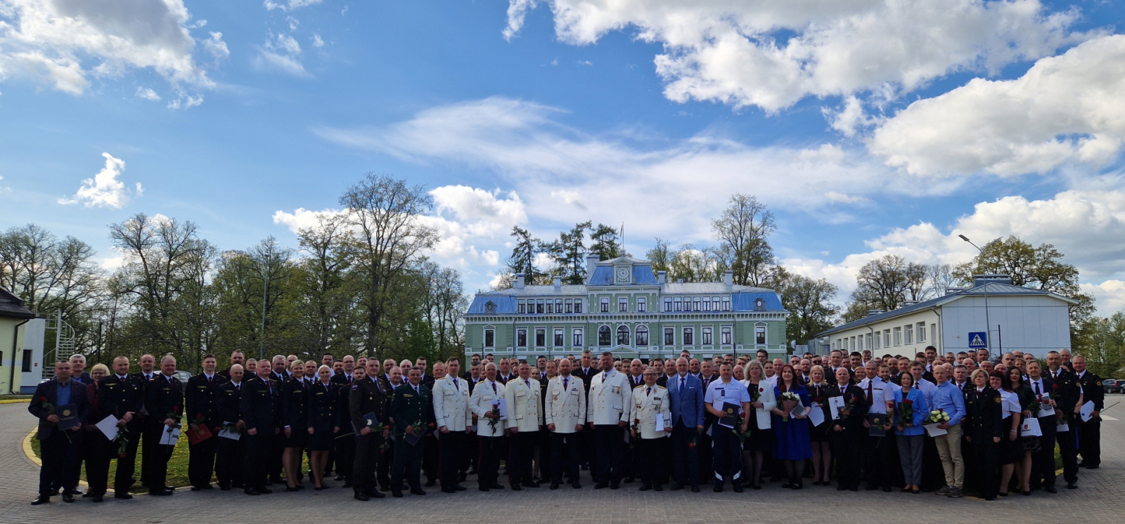 Formastērpos tērpušies VUGD nodarbinātie, kā arī viesi stāv un skatās fotogrāfa virzienā. Fonā gaiši zilas debesis ar baltiem mākoņiem un gaiša daudzstāvu ēka.