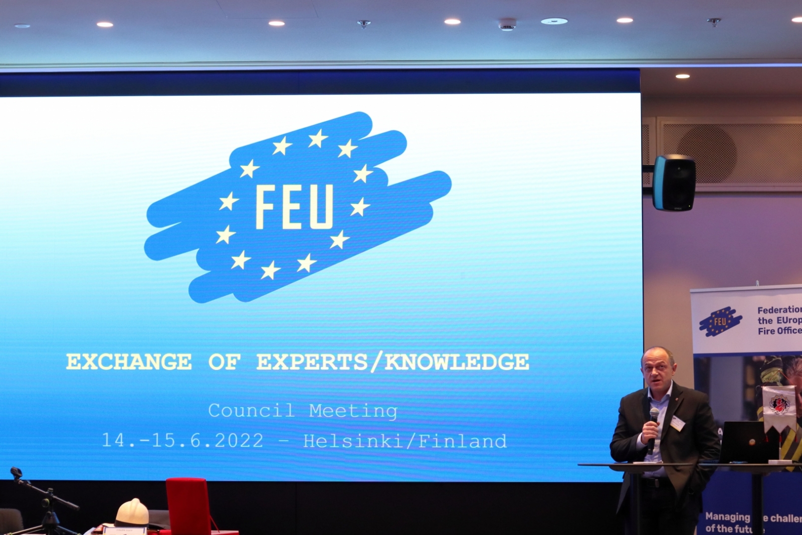 Auditorijas priekšā stāv vīrietis un rāda prezentāciju. Uz prezentācijas slaida redzams teksts angļu valodā "FEU. Exchagne of experts/ knowledge"