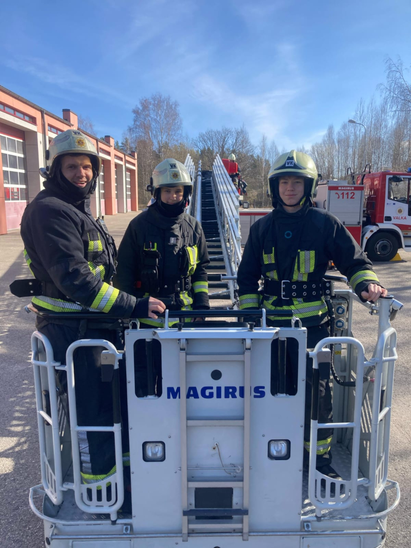 Pelēcīgās autokāpnēs stāv trīs ugunsdzēsēju glābēju aizsargtērpos un baltās aizsargķiverēs tērpti cilvēki.