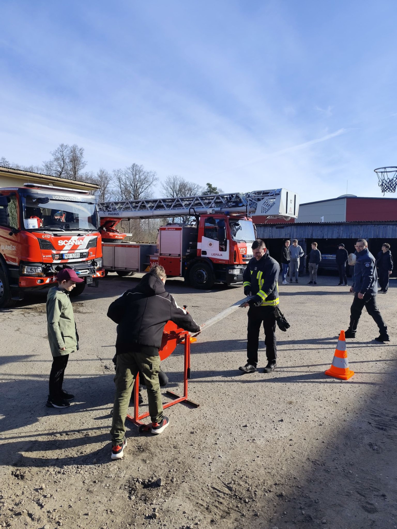 Pagalmā novietots sarkans instruments ar kura palīdzību jaunietis kopā ar ugunsdzēsēju glābēju tin ūdens šļūteni. Otrs jaunietis stāv blakus un skatās notiekošajā.