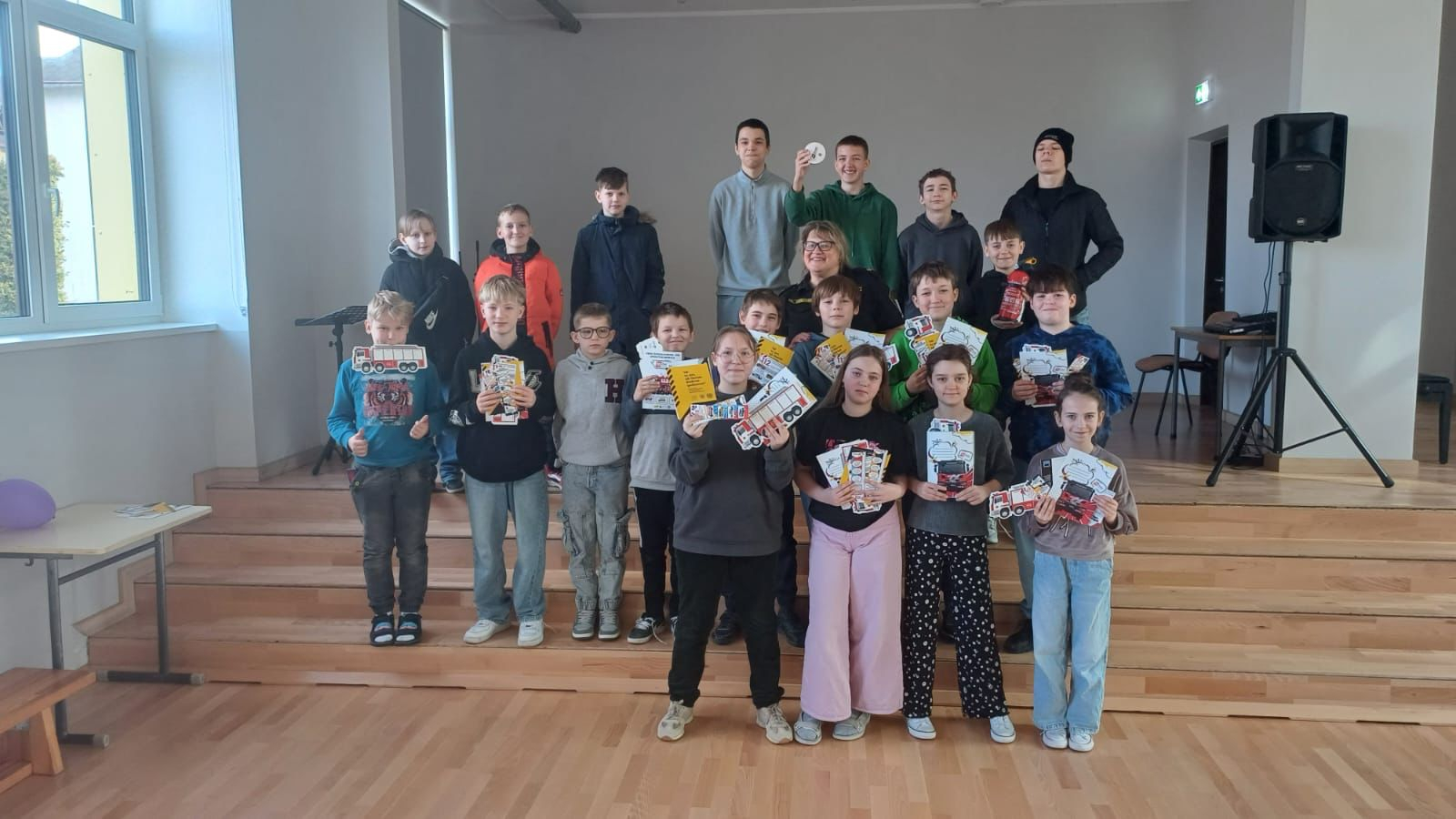Iekštelpās uz brūnām skatuves koka kāpnītēm nostājušies jaunieši kopā ar inspektori. Visi skatās fotogrāfa virzienā. Jaunieši rokās tur iegūtos informatīvos materiālus un reprezentācijas priekšmetus.