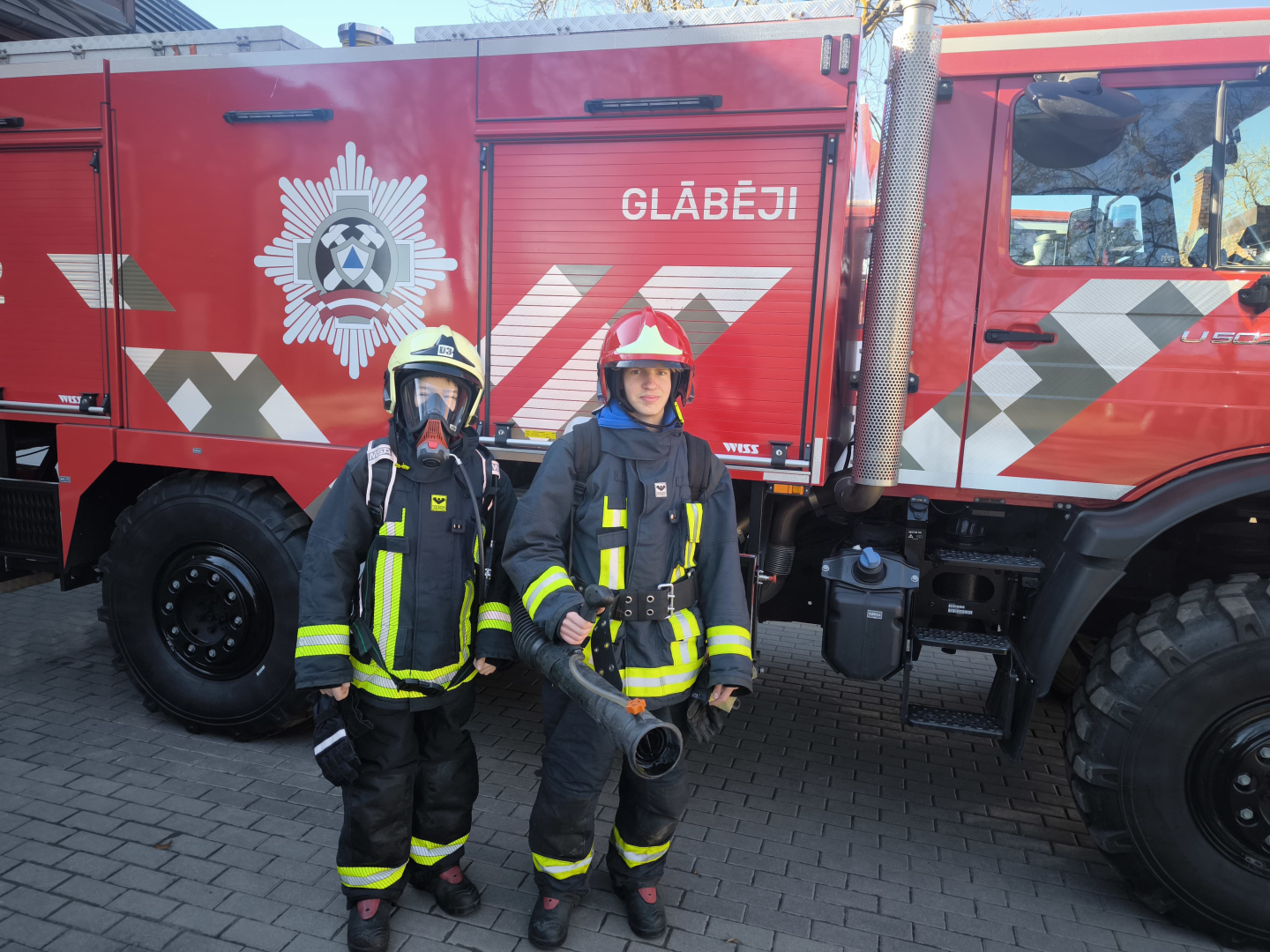 Pie ugunsdzēsēju glābēju autocisternas stāv divi jaunieši, kas tērpušies ugunsdzēsēju glābēju aizsargtērpos. Viens no jauniešiem ir uzvilcis aizsargmasku, savukārt otrs tur rokās vienu no ugunsdzēsēju glābēju darbarīkiem.