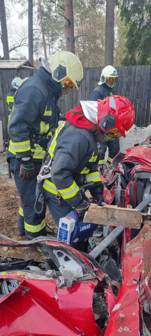 Jaunietis, kas tērpies ugunsdzēsēja glābēja aizsargtērpā un galvā uzlicis sarkanu aizsargķiveri stāv pieliecies pie sarkanas automašīnas un mēģina ar hidrauliskā instrumenta palīdzību atvērt tās durvis. Jaunietim blakus stāv ugunsdzēsējs glābējs, kurš vēro un stāsta par darbiem, kas tiek veikti ar hidrauliskajiem instrumentiem.