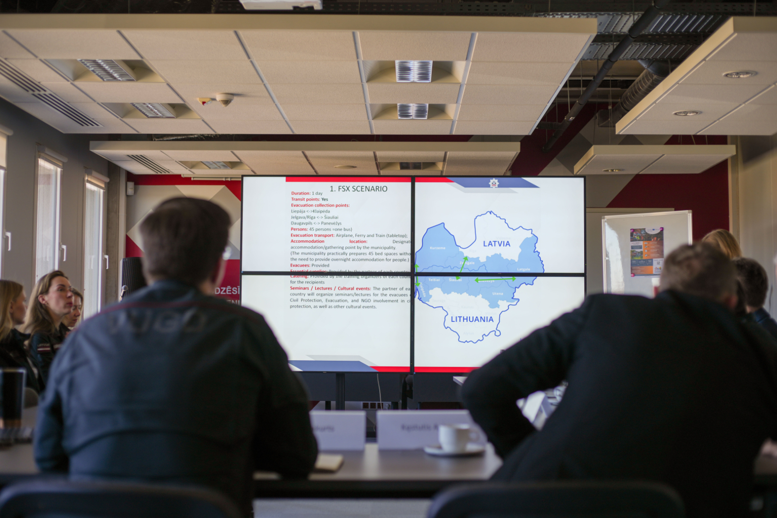 Sanāksmju zāle. Priekšplānā divi vīrieši ar mugurpusi. Skats uz ekrānu, kurā tiek rādīta projekta prezentācija.