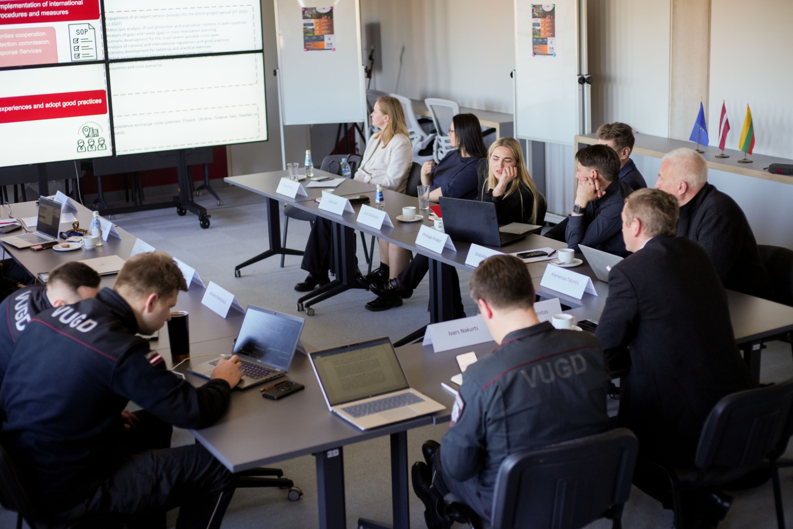 Sanāksmju zāle. Pie galda projekta pārstāvji. Pretī ekrāns ar prezentāciju.