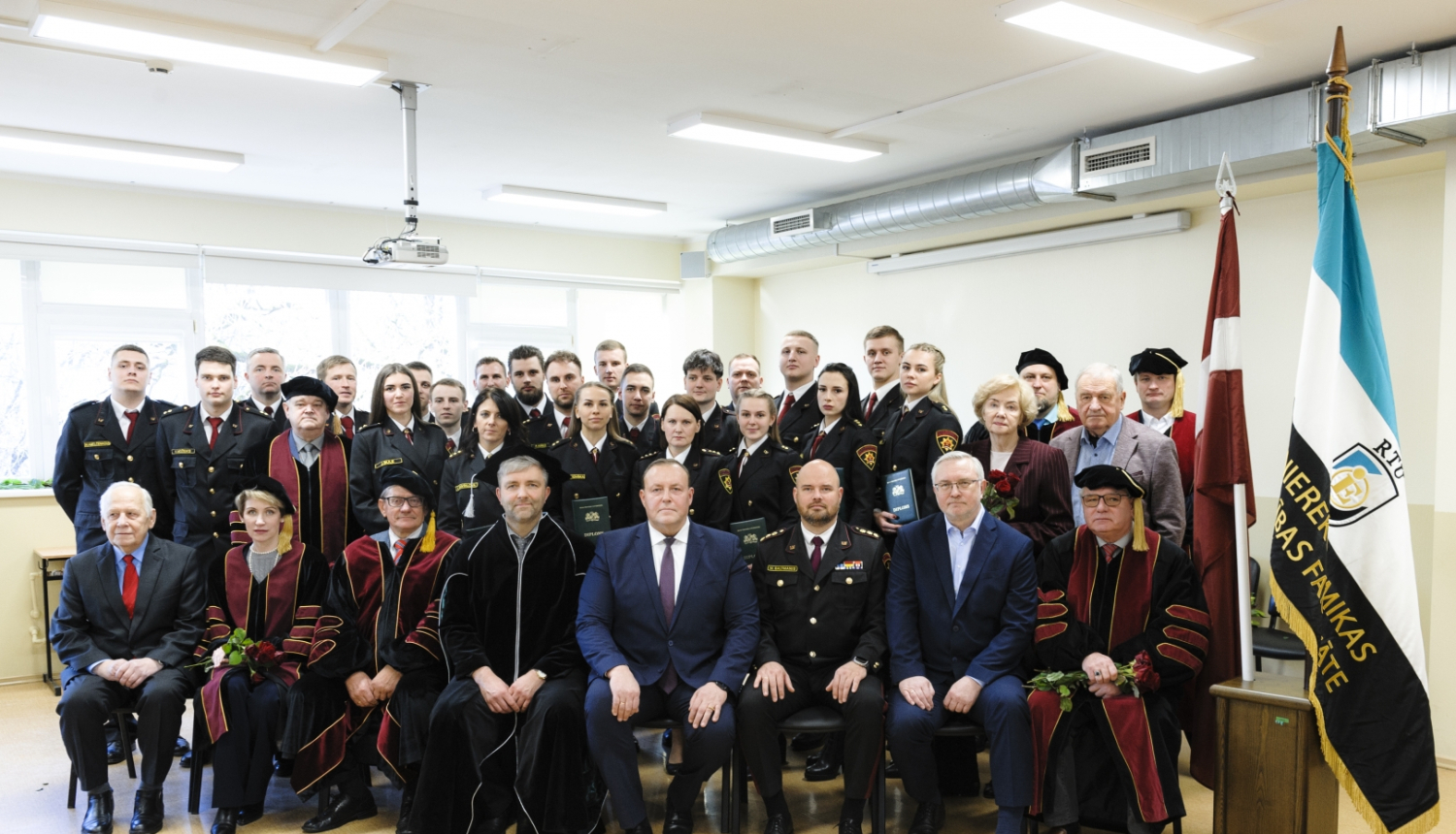 Izlaiduma dalībnieku kopbilde. Pirmajā rindā cilvēki sēž uz krēsliem, bet viņiem aiz muguras stāv citi izlaiduma dalībnieki un absolventi.