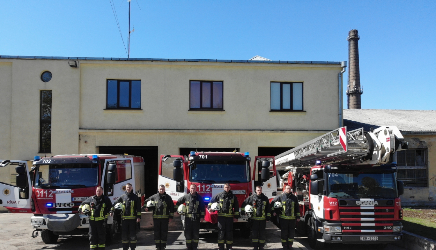 Septiņi ugunsdzēsēji glābēji stāv, turot rokās savas ķiveres. Viņiem fonā redzamas trīs dažādas ugunsdzēsēju glābēju autotehnikas