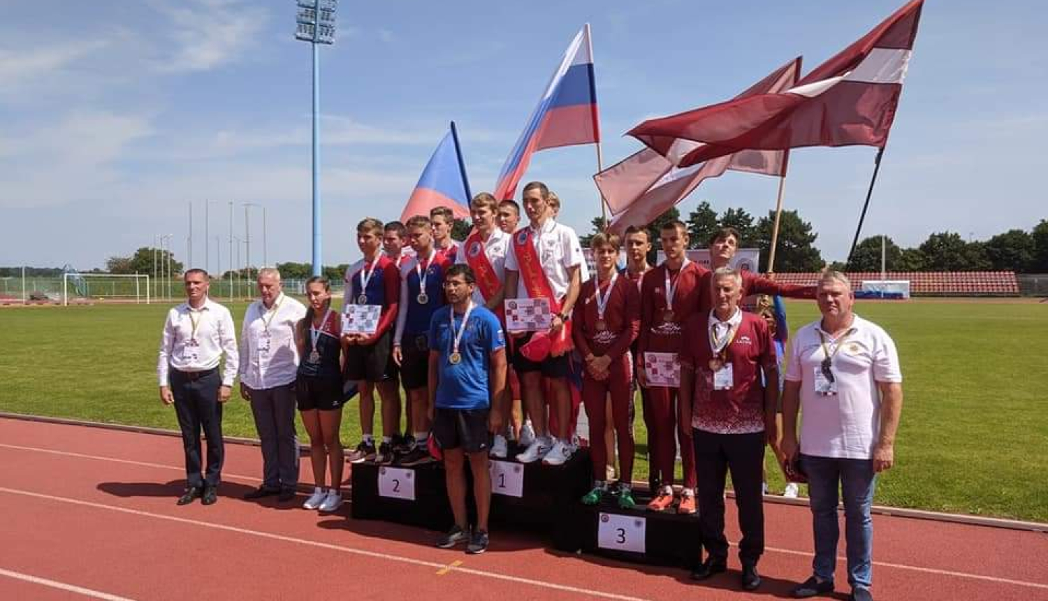 Uz stadiona sarkanīgā skrejceļa novietoti pjedestāli. Uz tiem uzkāpuši sacensību dalībnieki un uzvarētāji. Tiem aiz muguras novietoti trīs valstu karogi.