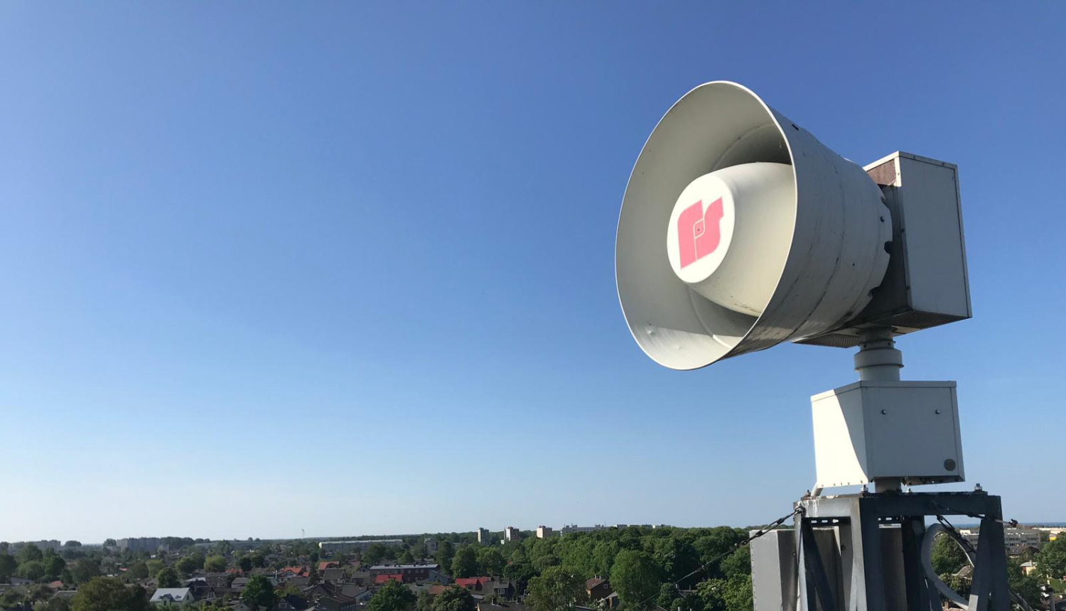 Uz jumta izvietota balta trauksmes sirēna. Tālumā redzamas zilas debesis, māju jumti un zaļas koku galotnes.