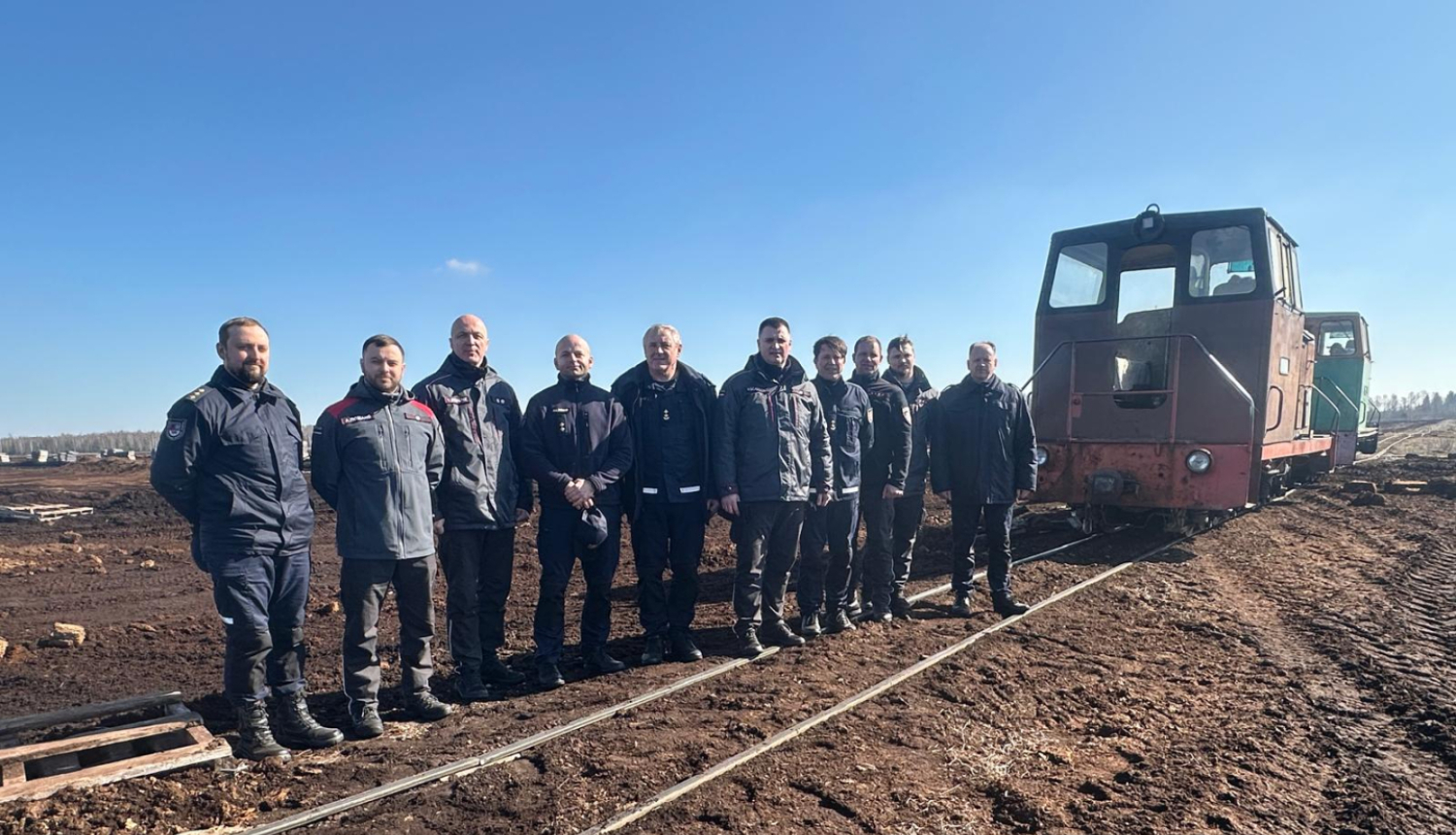 Pie kūdras laukā esošām vilciena sliedēm stāv operatīvo dienestu nodarbinātie un skatās fotogrāfa virzienā. Labajā malā redzams arī neliels kūdras laukā esošs vilcieniņš