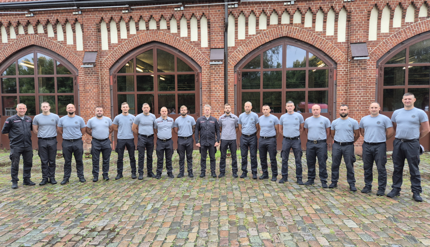 Septiņpadsmit VUGD pelēkajos formastērpos ģērbušies vīrieši stāv uz bruģa un skatās fotogrāfa virzienā. Aiz viņiem atrodas sarkana ķieģeļu māja ar lieliem stiklotiem arkveida logiem.