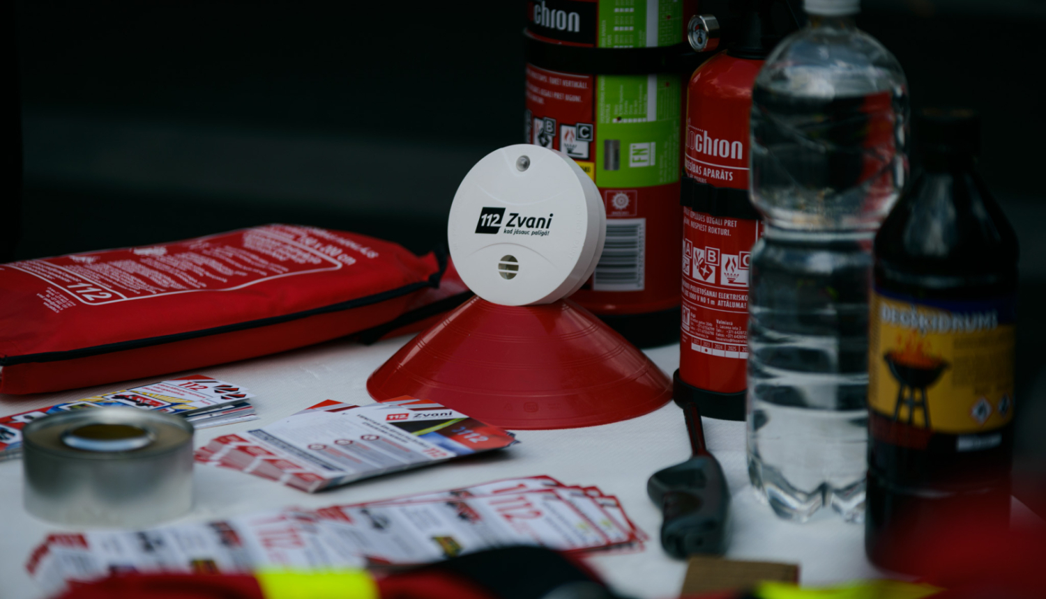 Uz galda novietoti dažādi priekšmeti - ugunsdzēsības pārklājs, statīvs uz kura izvietots balts dūmu detektors, bet uz tā melniem cipariem un burtiem rakstīts: 112 Zvani, kad jāsauc palīgā. Vēl uz galda izvietoti divi ugunsdzēsības aparāti, dažādi dedzināšanas šķidrumi, sarkanas glābšanas vestes, skrejlapas par vienoto ārkārtas palīdzības izsaukumu numuru 112.