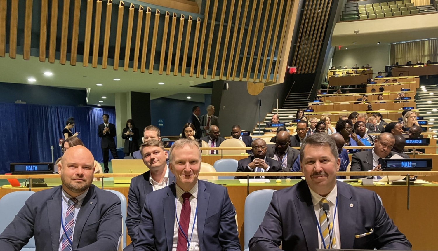 Trīs vīrieši, ģērbušies uzvalkos sēž pie koka galda ar zaļu pārklāju un skatās fotogrāfa virzienā. Uz malas novietots elektroniska ierīce uz kuras ziliem burtiem ir uzraksts - Latvia