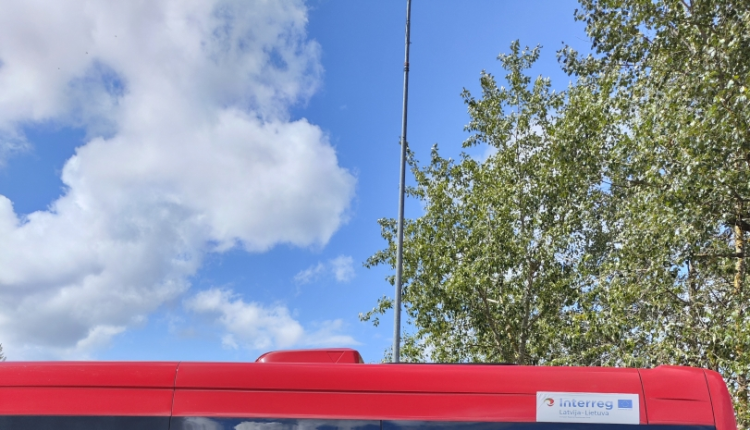 Sacensību uzvarētāju kopbilde. Visi cilvēki tērpušies melnās sporta drēbēs. Divi vīrieši un viena sieviete tur rokās katrs savu izcīnīto zelta kausu. Blakus esošiem sacensību dalībniekiem ap kaklu apliktas iegūtās medaļas. Fonā sarkanas krāsas mikroautobuss