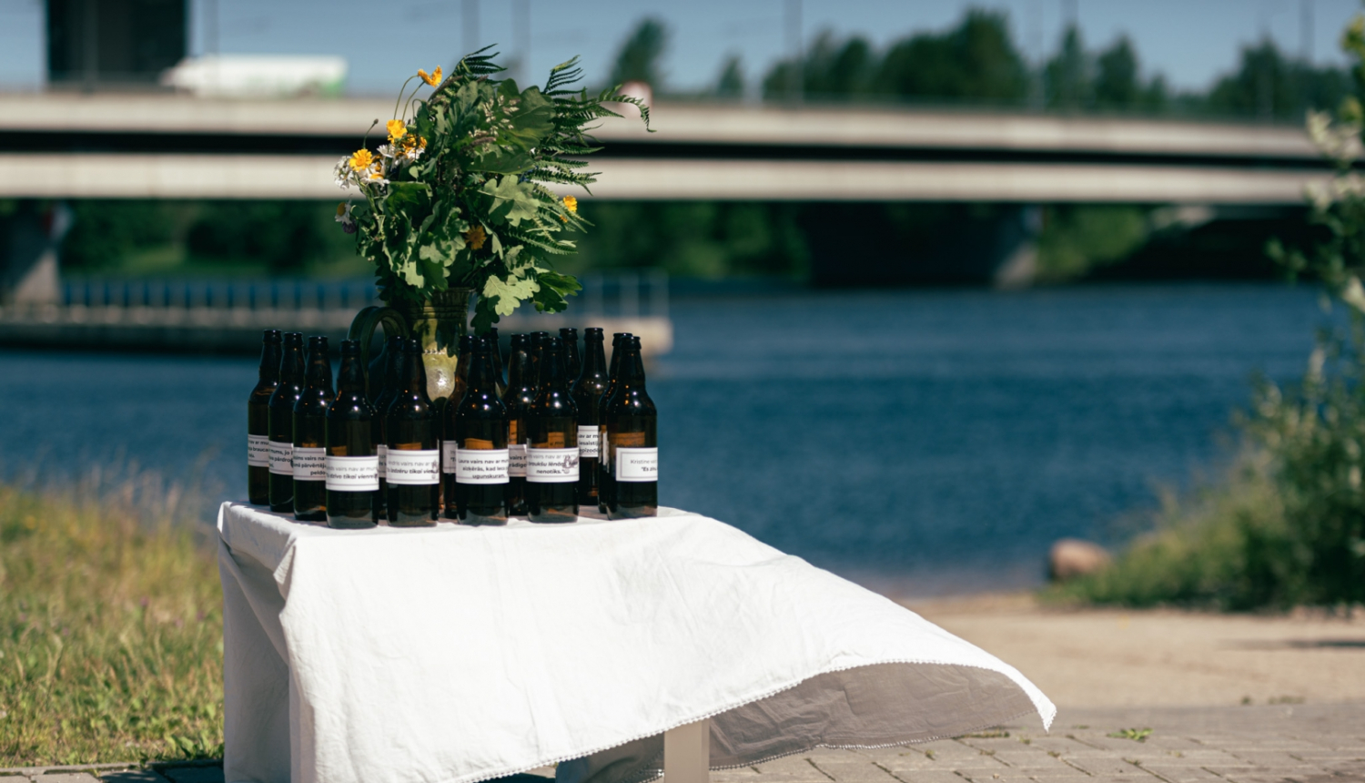 Uz maza galdiņa, kas apklāts ar baltu galdautu, novietotas tumši brūnas atvērtas alus pudeles un jāņuzāļu pušķis. Alus pudelēm uzlīmētas etiķetes ar aicinājumu aizdomāties par to, lai svētkus pavadītu droši