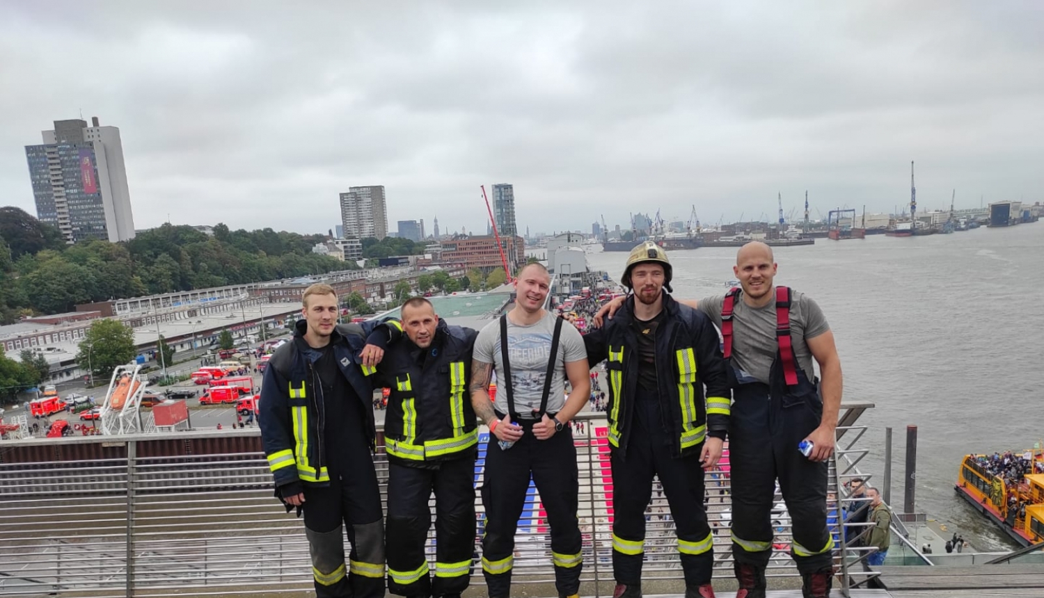 VUGD sportistu - piecu vīriešu - kopbilde. Trīs vīrieši tērpušies ugunsdzēsēju glābēju drēbēs.