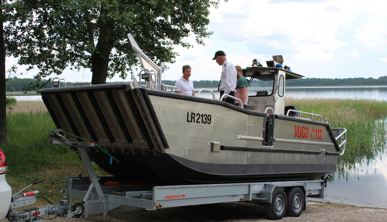 Ūdenstilpes krastā uz piekabes novietota metāla laiva. Laivā atrodas divi vīrieši.