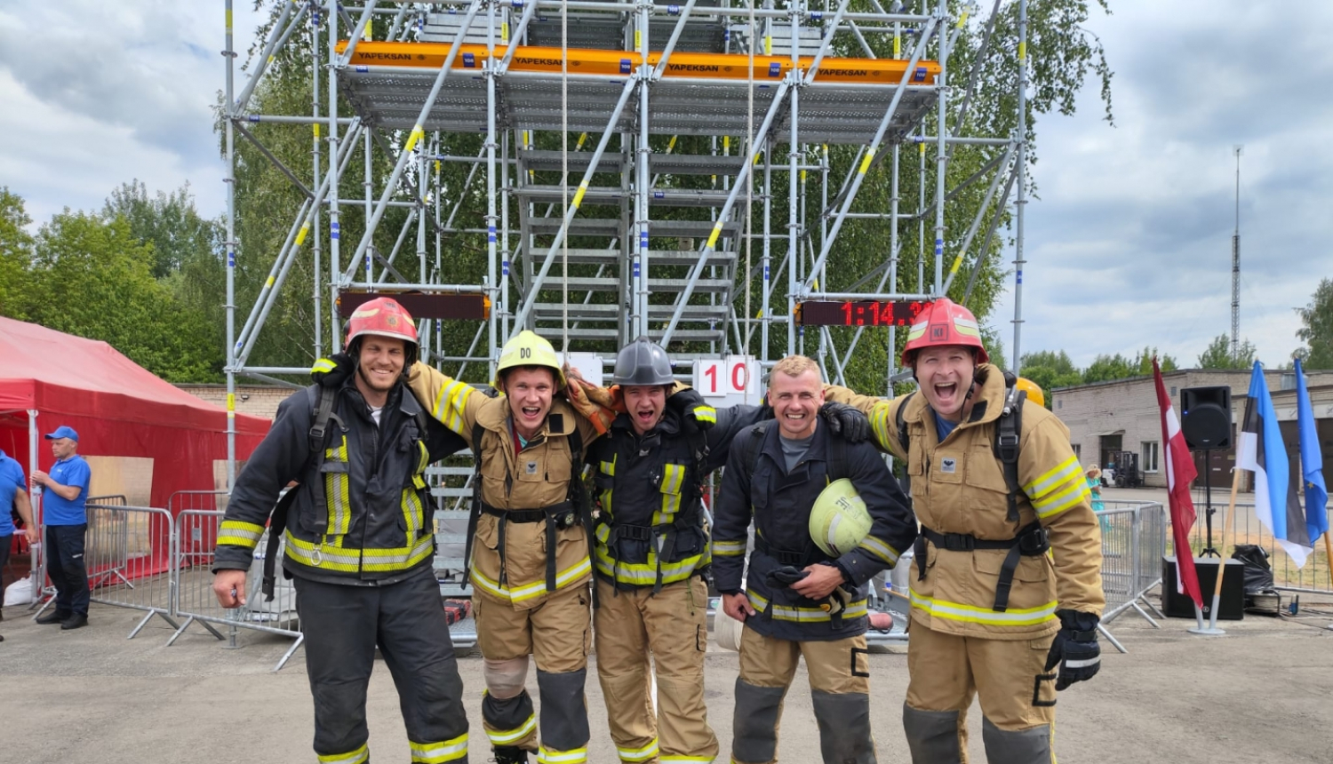 Pieci ugunsdzēsēji glābēji, tērpušies savos aizsagtērpos, stāv vienā rindā un skatās fotogrāfa virzienā. Fonā metāla torņa kāpnes