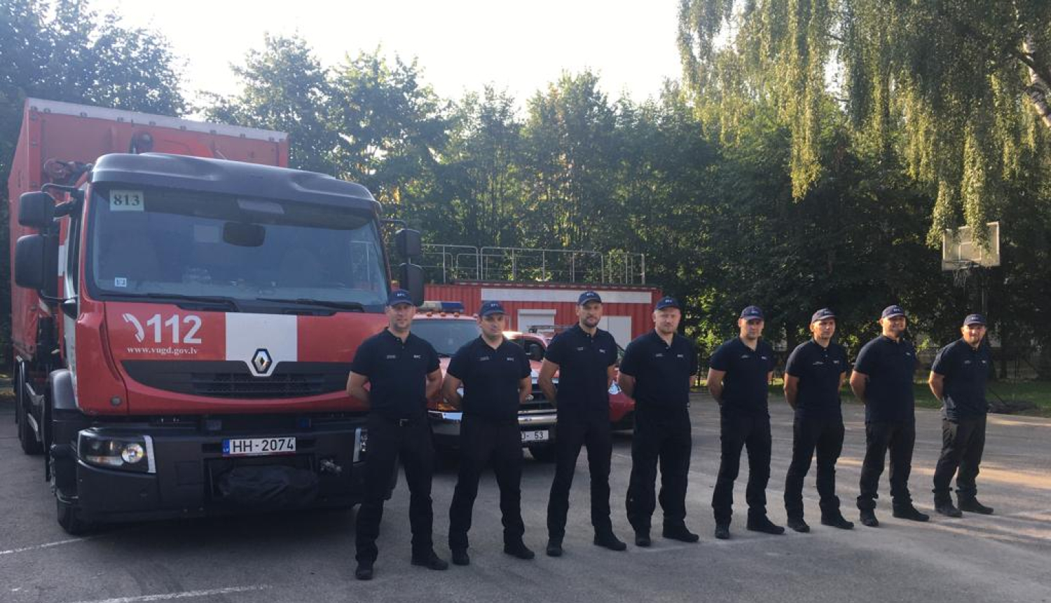 Astoņi vīrieši, tērpušies formastērpos, stāv vienā taisnā līnijā viens otram blakus un skatās fotogrāfa virzienā. Aiz viņiem novietoti VUGD sarkanbaltie transportlīdzekļi