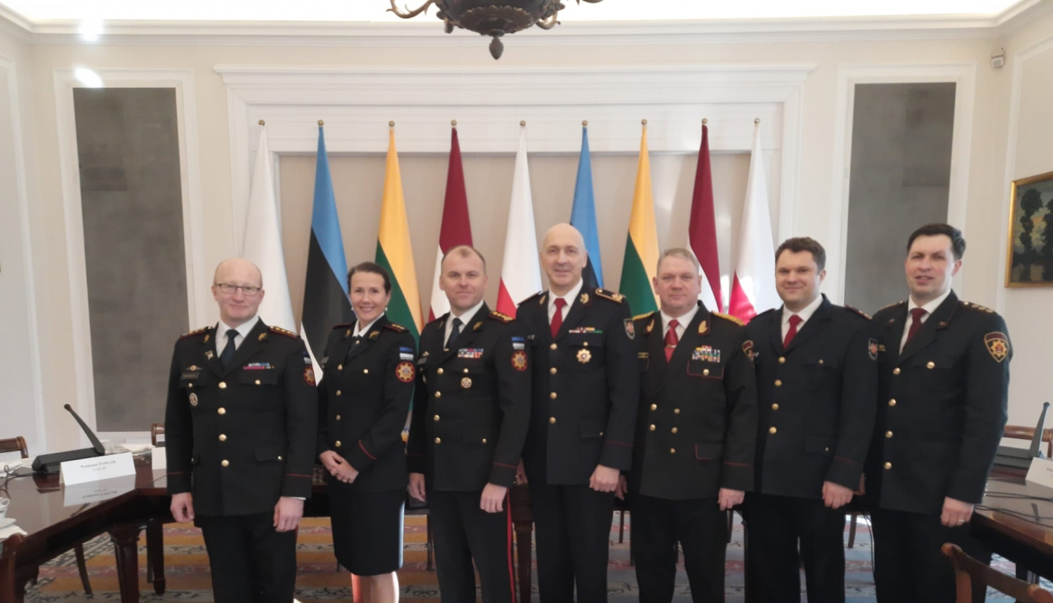 Septiņi konferences dalībnieki, tērpušies savās dienestu formās, stāv pie konferences dalībvalstu karogiem un skatās fotogrāfa virzienā