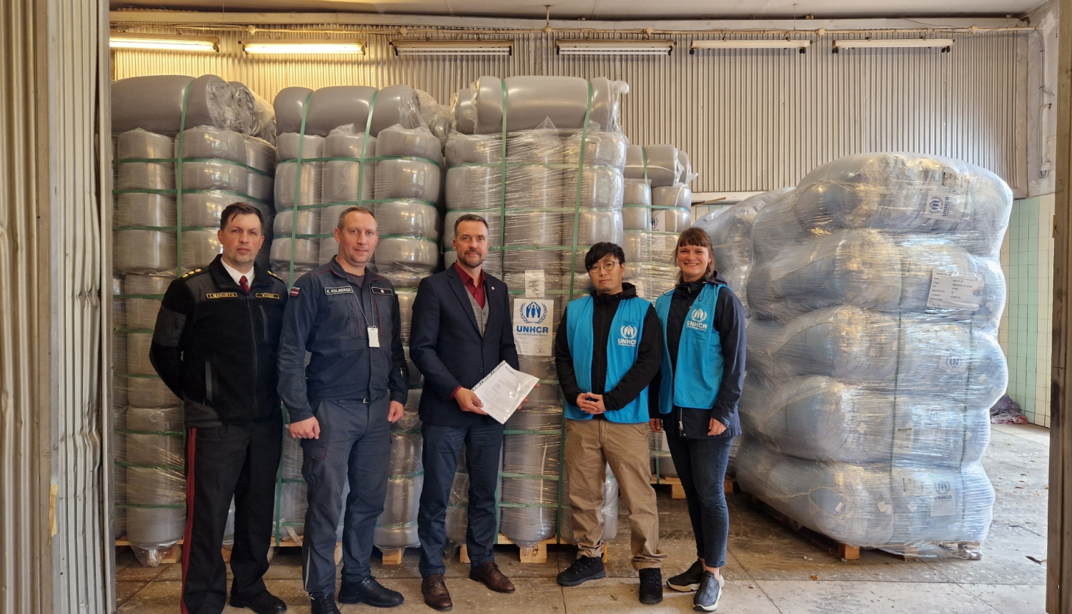 Divi VUGD darbinieki, Iekšlietu ministrs un divas ANO pārstāves stāv un skatās fotogrāfa virzienā. Fonā redzamas piegādātās mantas