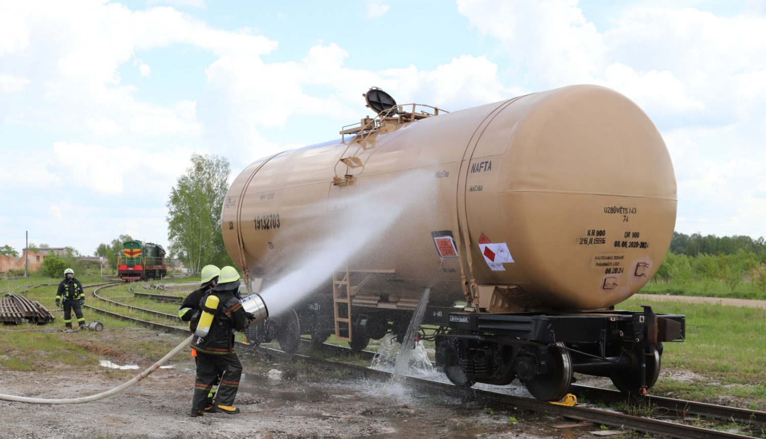 Uz dzelzceļa sliedēm stāv dzeltena cisterna. Tai blakus atrodas divi ugunsdzēsēji glābēji, kas uz cisternas ar šļūtenēm lej ūdeni