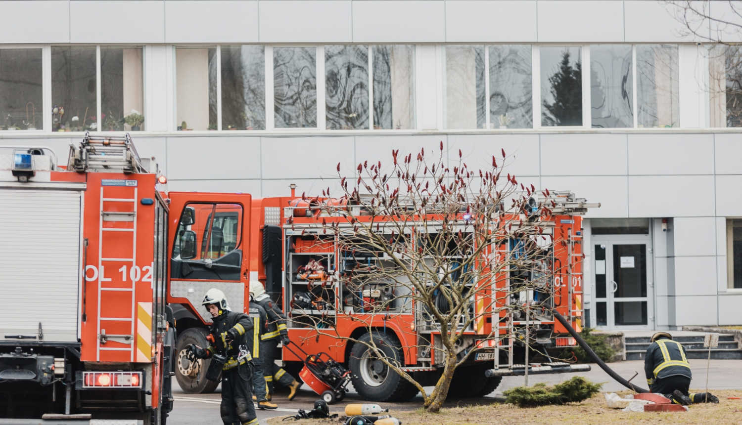Divas Valsts ugunsdzēsības un glābšanas dienesta mašīnas stāv pie baltas ēkas. Ap mašīnām rosās ugunsdzēsēji glābēji.