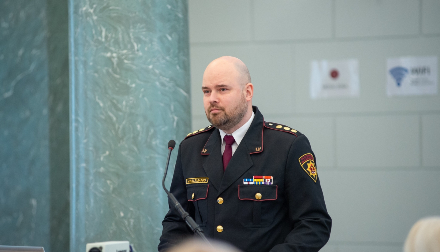 Vīrietis, tērpies dienesta formastērpā, stāv pie galdiņa ar mikrofonu auditorijas priekšā. 