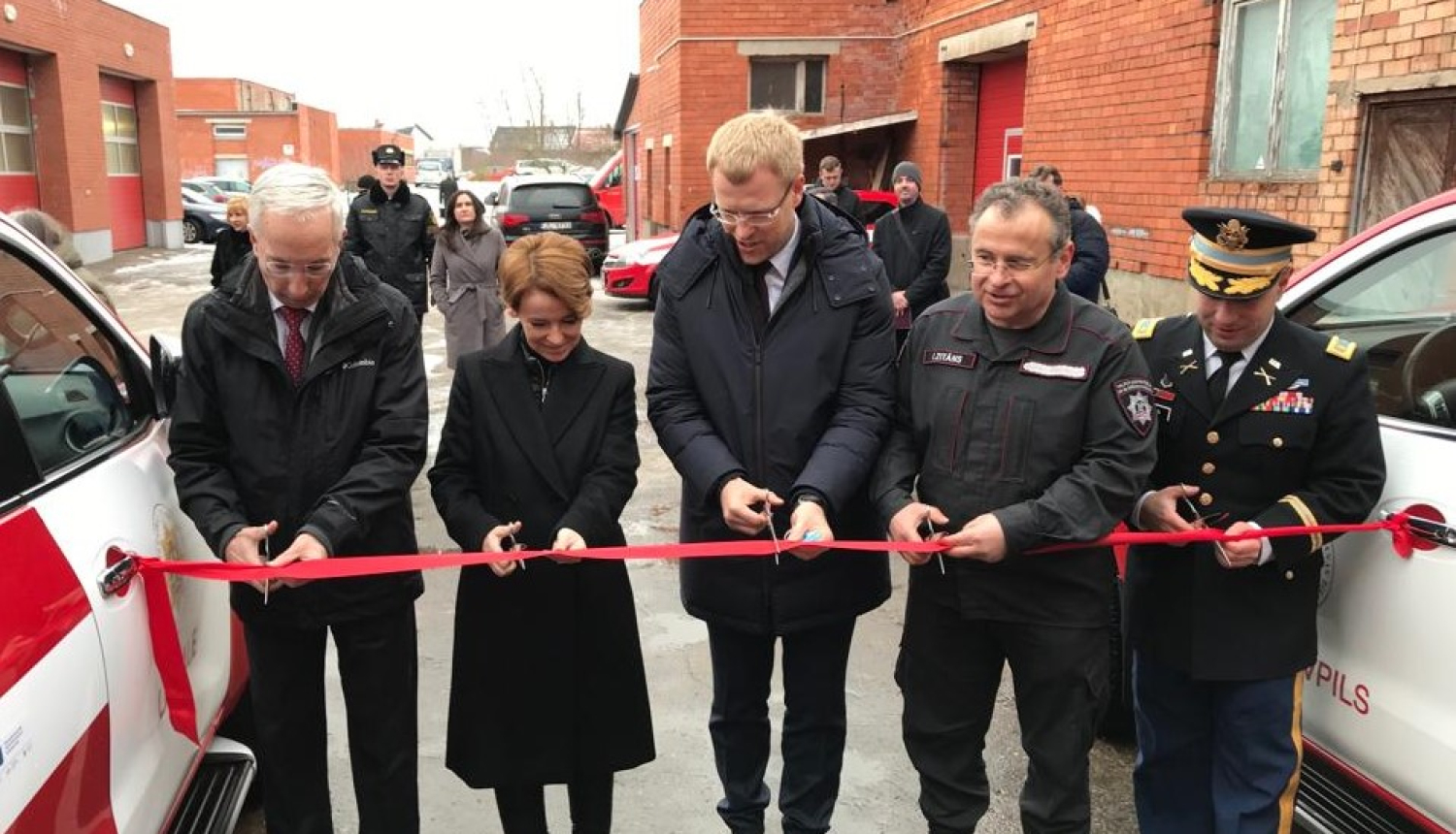 Pieci cilvēki stāv pie sarkanas lentes, kas novilkta no viena ugunsdzēsēju glābēju operatīvā transportlīdzekļa uz otru. Cilvēkiem rokās šķēres. Viņi grasās pārgriezt sarkano lenti.
