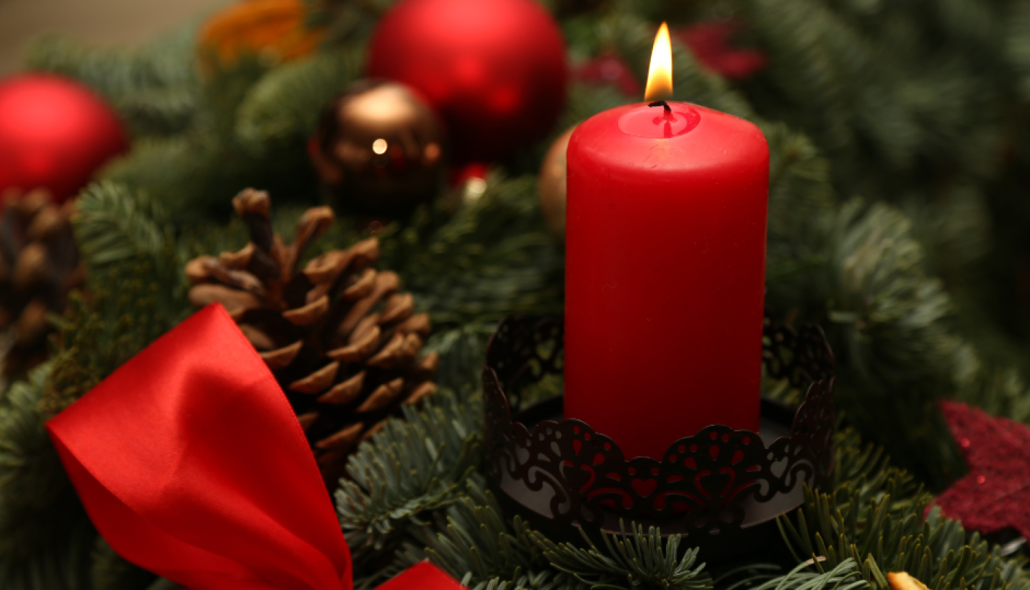 Adventes vainagā aizdegta viena sarkana svece. Netālu no tās atrodas brūns čiekurs, sarkana bantīte, kā arī citi dekoratīvie elementi.