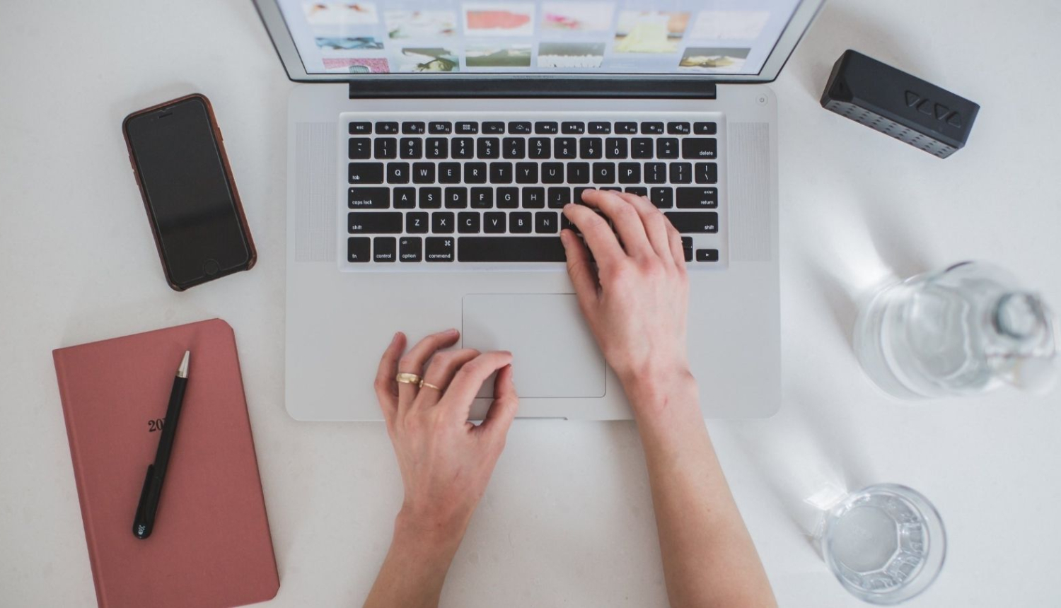 Uz balta galda novietots atvērts portatīvais dators. Tā cilvēks ar savu labo roku pieskaras klaviatūrai, savukārt kreisā roka novietota uz galda. Datora kreisajā malā novietots mobilais tālrunis un rozīgas krāsas blociņš uz kura novietota pildspalva. Datoram labajā malā novietota stikla ūdens pudele un glāze. 