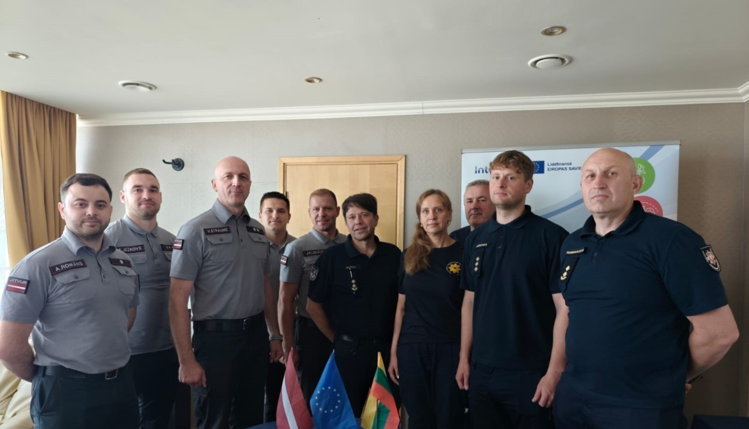 Desmit Latvijas un Lietuvas operatīvo dienestu nodarbinātie stāv ap mazu galdiņu uz kura novietoti trīs karogi - Latvijas, Igaunijas un Lietuvas