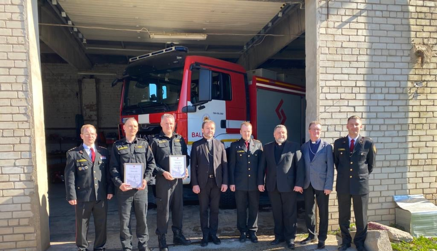 Pie ēkas ar vaļējām durvīm stāv astoņi vīrieši - ugunsdzēsēji glābēji un Baldones baznīcas pārstāvji. Fonā redzama ugunsdzēsēju glābēju automašīna