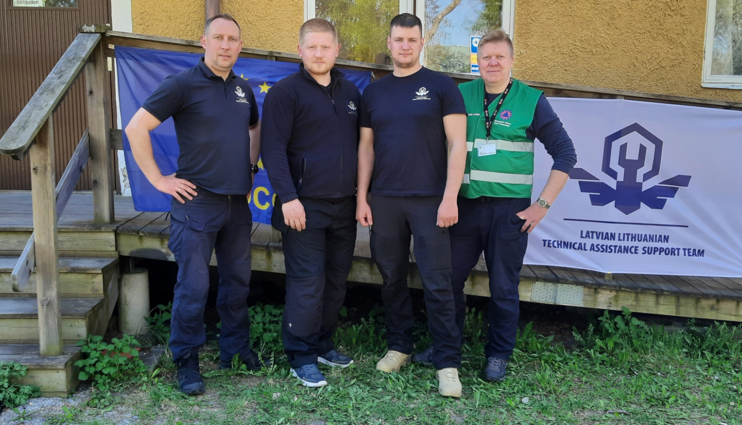 Četri vīrieši stāv zālienā. Fonā redzama dzeltena ēkas siena, kāpnes