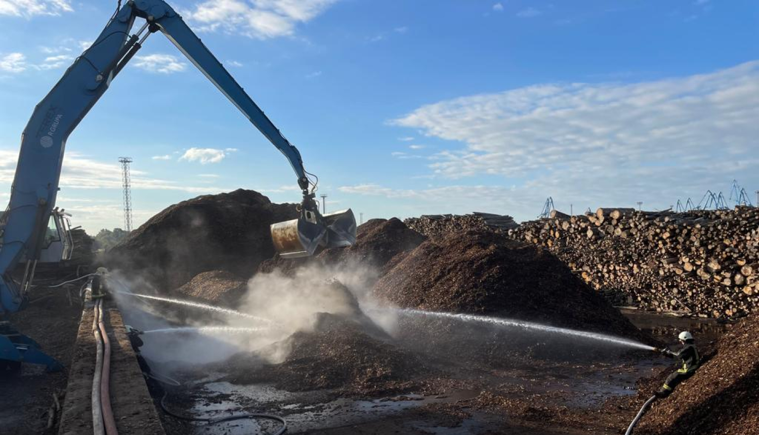 Liels eskavators virs šķeldas kaudzes tur savus kausus. No trīs pusēm ugunsdzēsēji glābēji dzēš gruzdošo šķeldu. Fonā zilas debesis ar baltiem mākonīšiem