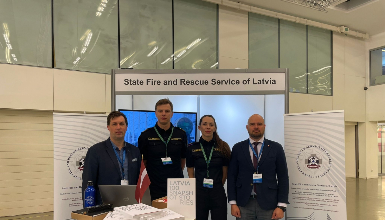 Četri VUGD darbinieki - trīs vīrieši un viena sieviete stāv pie galdiņa uz kura izvietoti dažādi uzskates materiāli. Fonā redzami divi baneri.