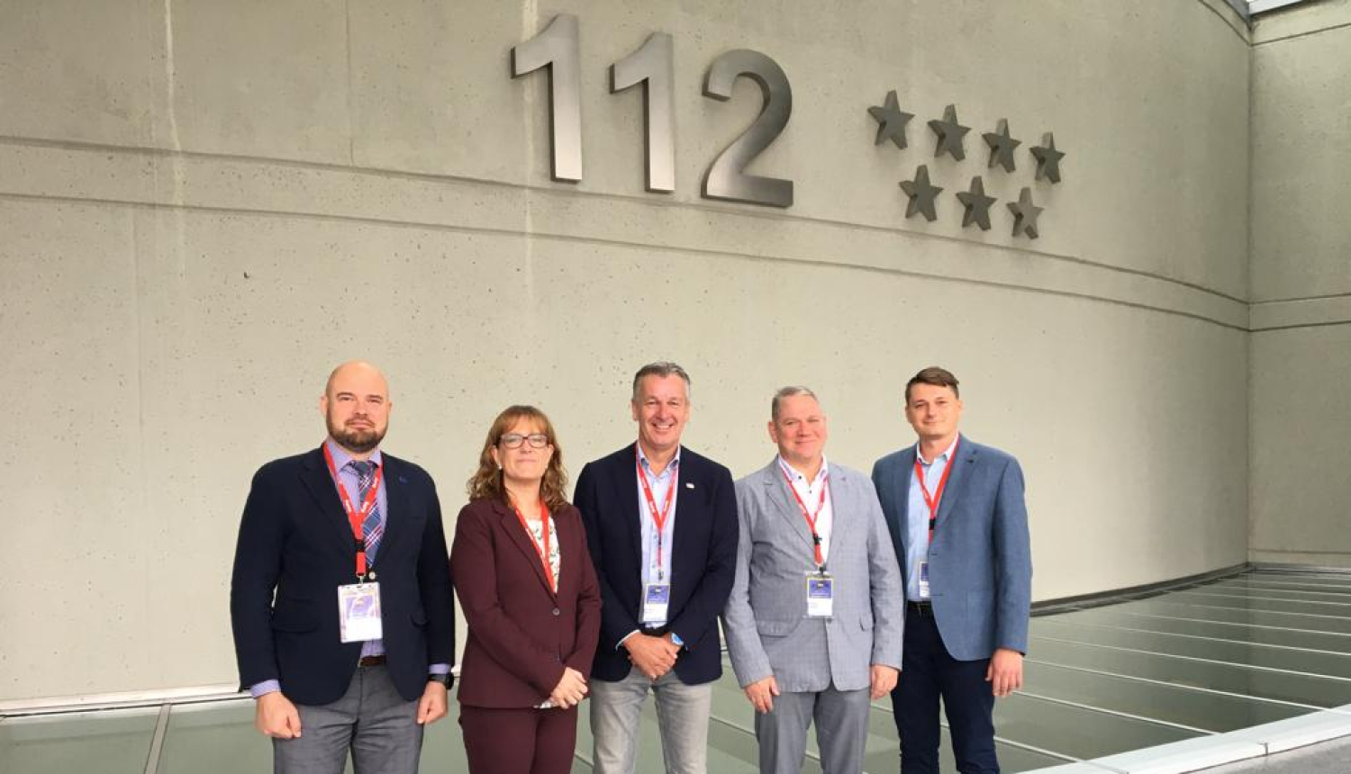 Piecu cilvēku - vienas sievietes un četru vīriešu - kopbilde. Visi stāv un skatās fotogrāfa virzienā. Fonā redzama siena uz kuras ir redzams tālruņa numurs 112 un septiņas zvaigznes.