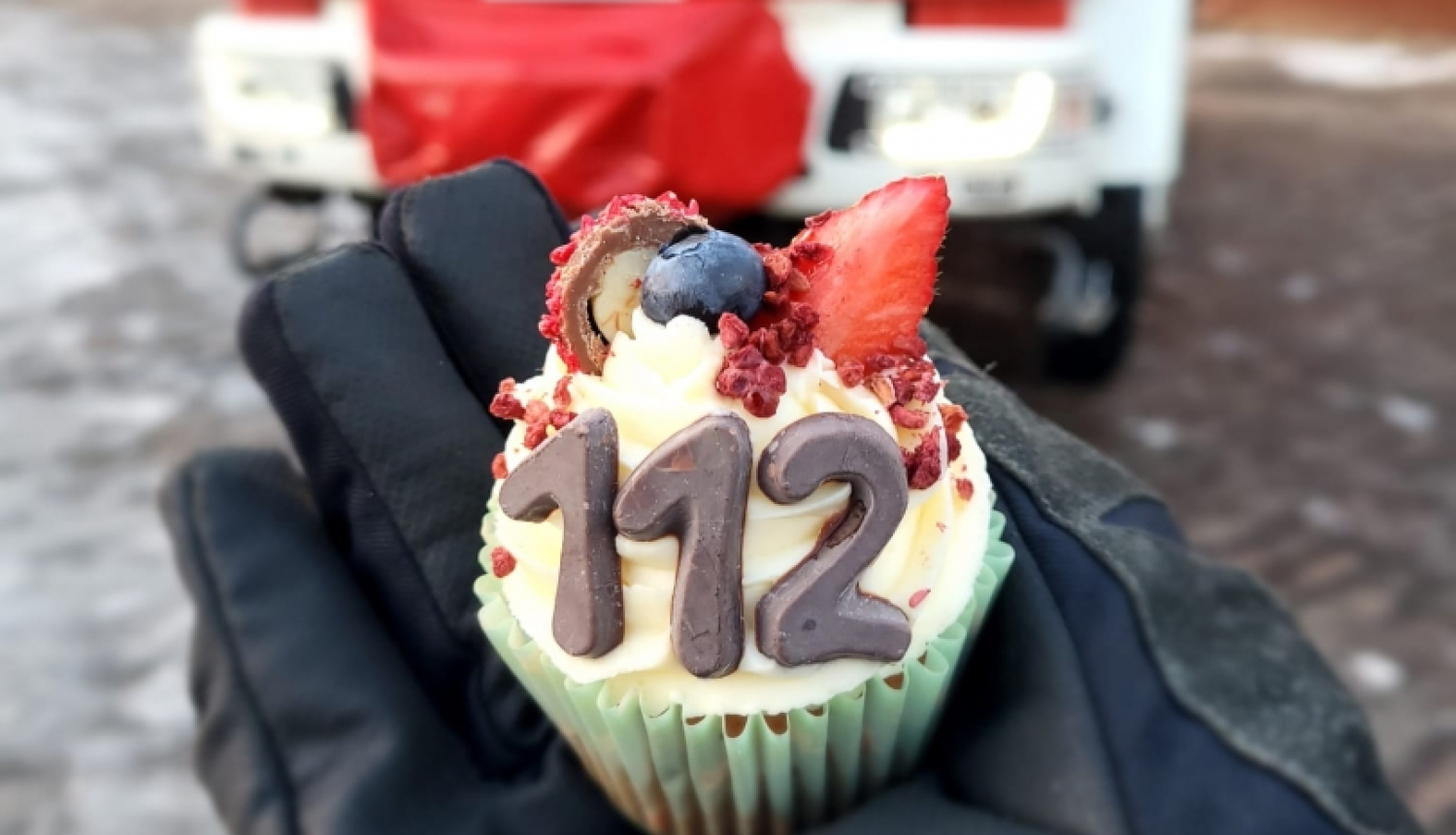 Redzamas cilvēka plaukstas, kas ietērptas melnos cimdos. Tajās tiek turēts mazs konditorejas izstrādājums ar cipariem 112. Fonā novietota ugunsdzēsēju glābēju autocisterna