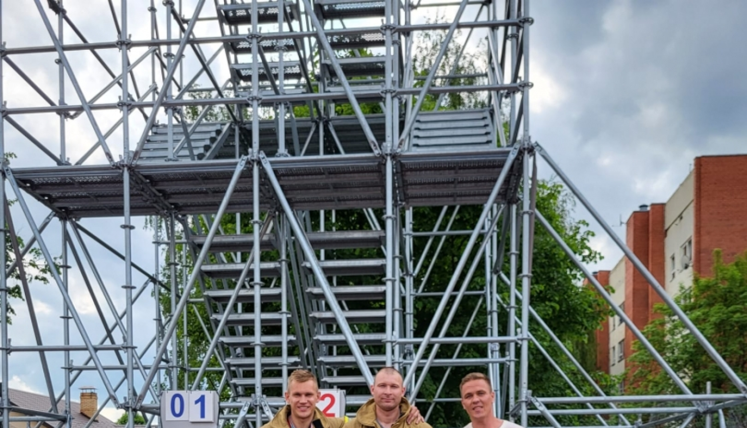 Uz pjedestāla stāv trīs vīrieši. Divi no tiem tērpušies bēšīgas krāsas formastērpos. Visi vīrieši rokās tur savas iegūtās balvas. Viņiem fonā redzamas metāla torņa kāpnes.