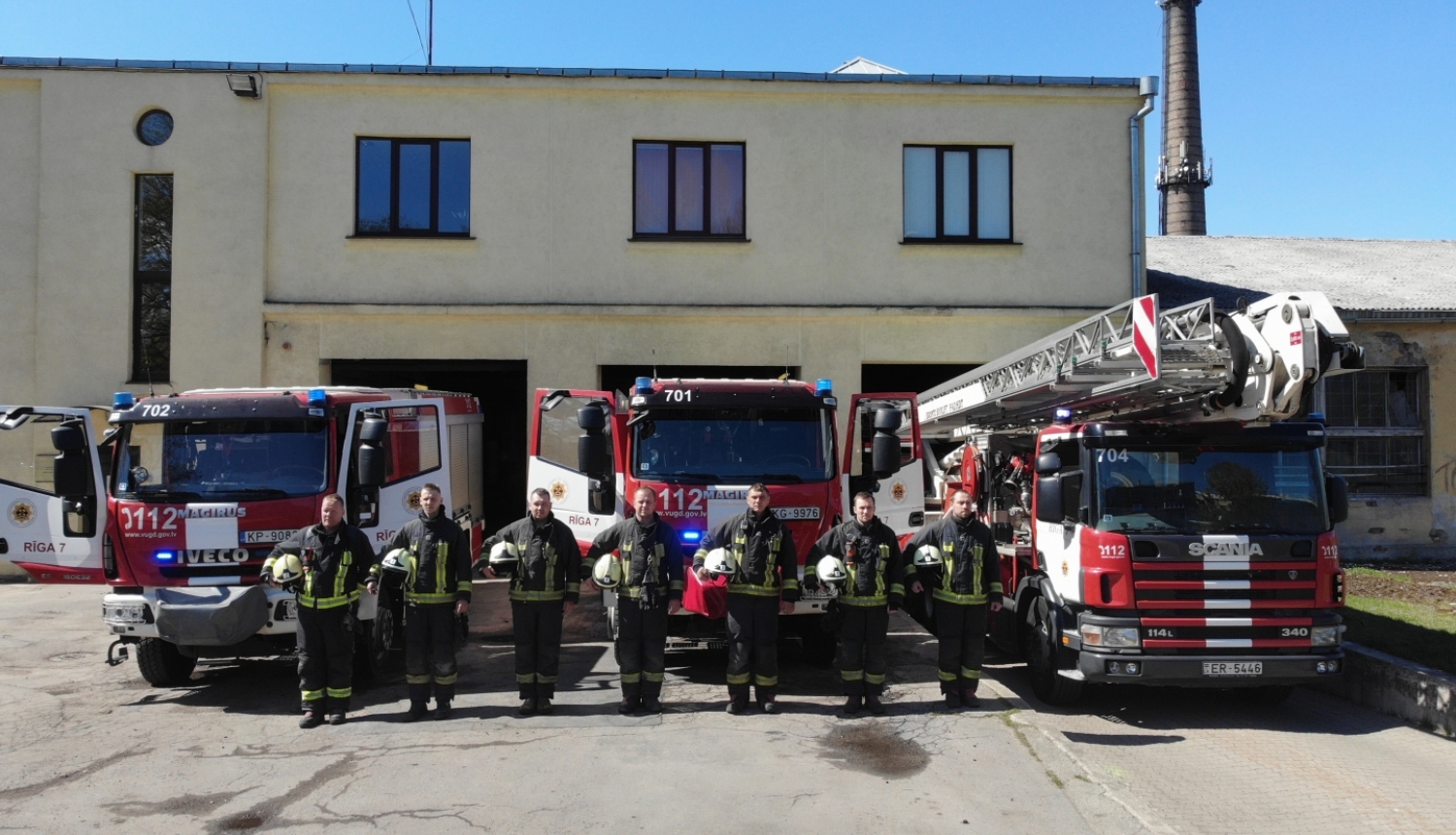 Septiņi ugunsdzēsēji glābēji stāv, turot rokās savas ķiveres. Viņiem fonā redzamas trīs dažādas ugunsdzēsēju glābēju autotehnikas