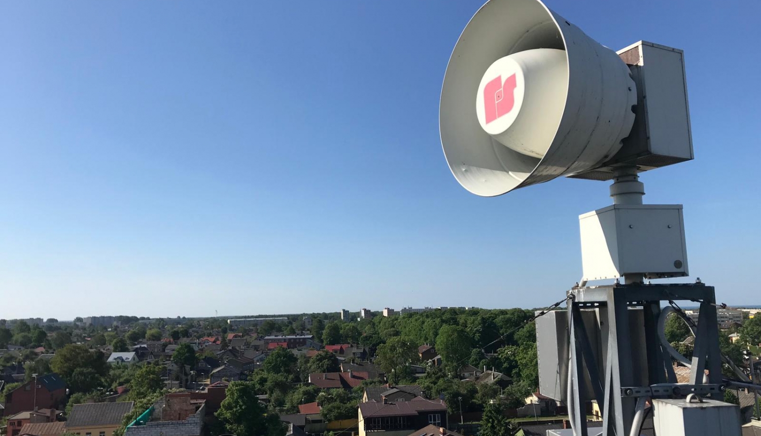 Uz jumta izvietota balta trauksmes sirēna. Tālumā redzamas zilas debesis, māju jumti un zaļas koku galotnes.