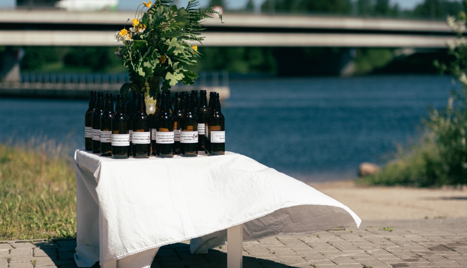 Uz maza galdiņa, kas apklāts ar baltu galdautu, novietotas tumši brūnas atvērtas alus pudeles un jāņuzāļu pušķis. Alus pudelēm uzlīmētas etiķetes ar aicinājumu aizdomāties par to, lai svētkus pavadītu droši