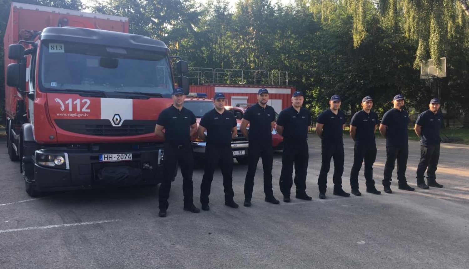 Astoņi vīrieši, tērpušies formastērpos, stāv vienā taisnā līnijā viens otram blakus un skatās fotogrāfa virzienā. Aiz viņiem novietoti VUGD sarkanbaltie transportlīdzekļi