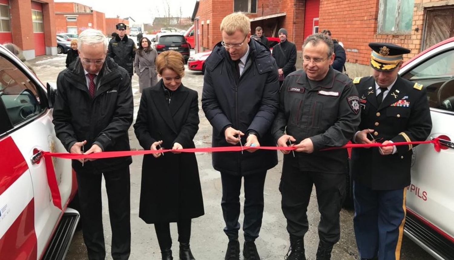 Pieci cilvēki stāv pie sarkanas lentes, kas novilkta no viena ugunsdzēsēju glābēju operatīvā transportlīdzekļa uz otru. Cilvēkiem rokās šķēres. Viņi grasās pārgriezt sarkano lenti.