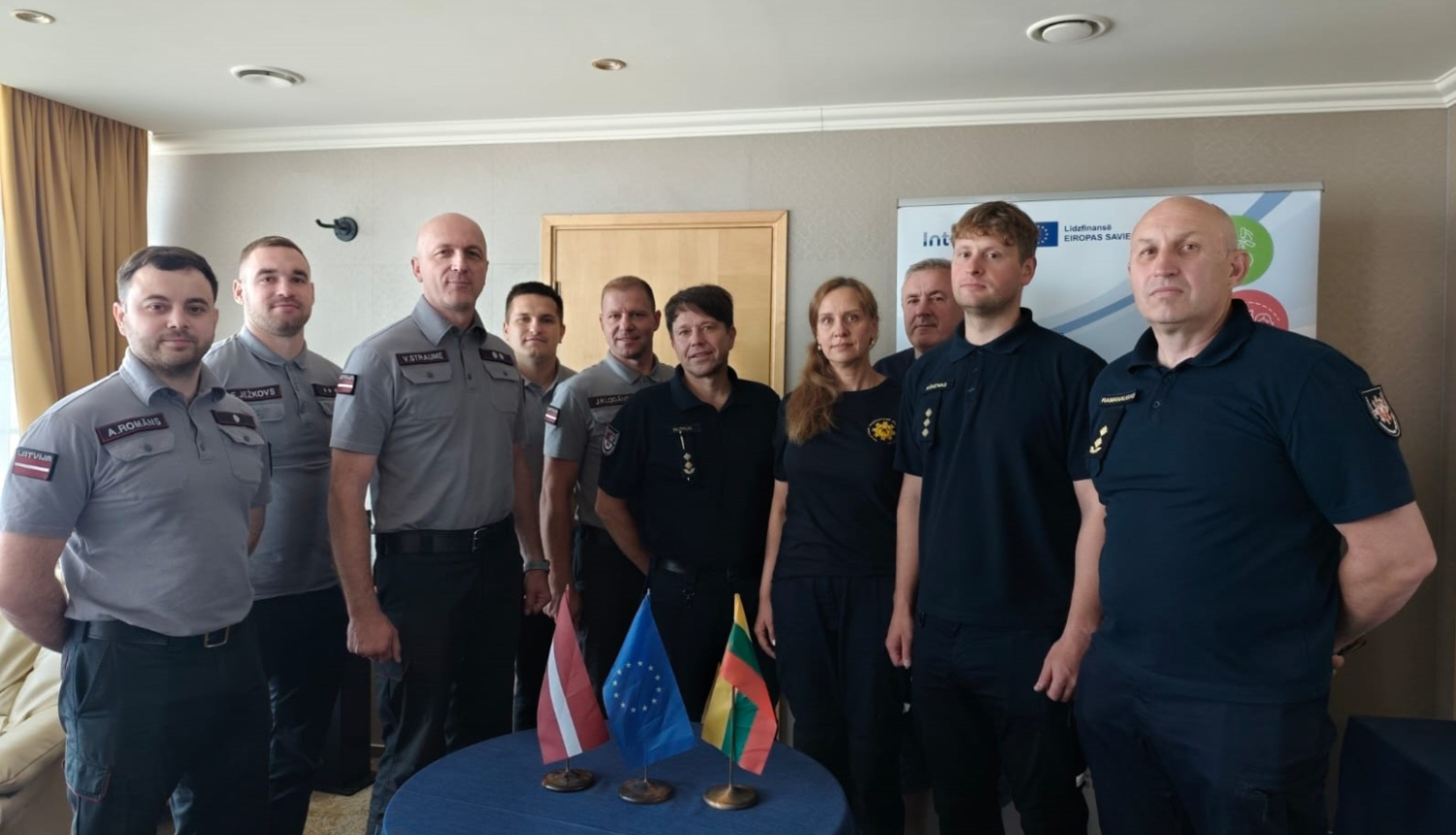 Desmit Latvijas un Lietuvas operatīvo dienestu nodarbinātie stāv ap mazu galdiņu uz kura novietoti trīs karogi - Latvijas, Igaunijas un Lietuvas