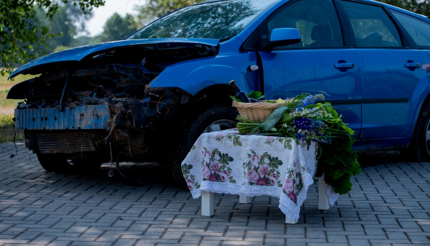 Stāv zila, sasista automašīna. Tai priekšā uz balta galdiņa, kas pārklāts ar raibu galdautu, uzliktas jāņuzāles un grozs ar alkoholu, kā arī svētku gardumiem