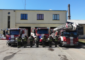 Septiņi ugunsdzēsēji glābēji stāv, turot rokās savas ķiveres. Viņiem fonā redzamas trīs dažādas ugunsdzēsēju glābēju autotehnikas